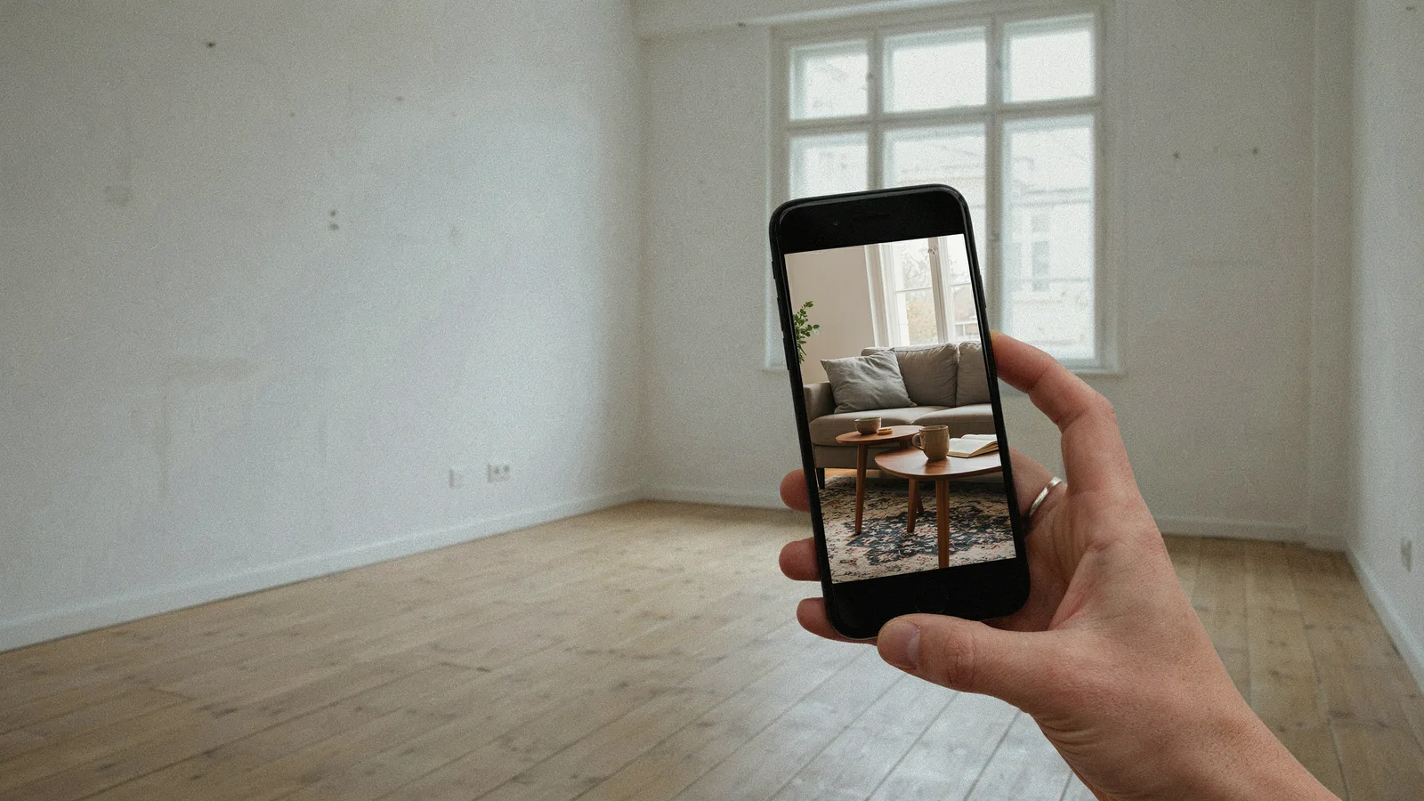 A phone showing a virtually staged living room, held up in front of the same room standing empty
